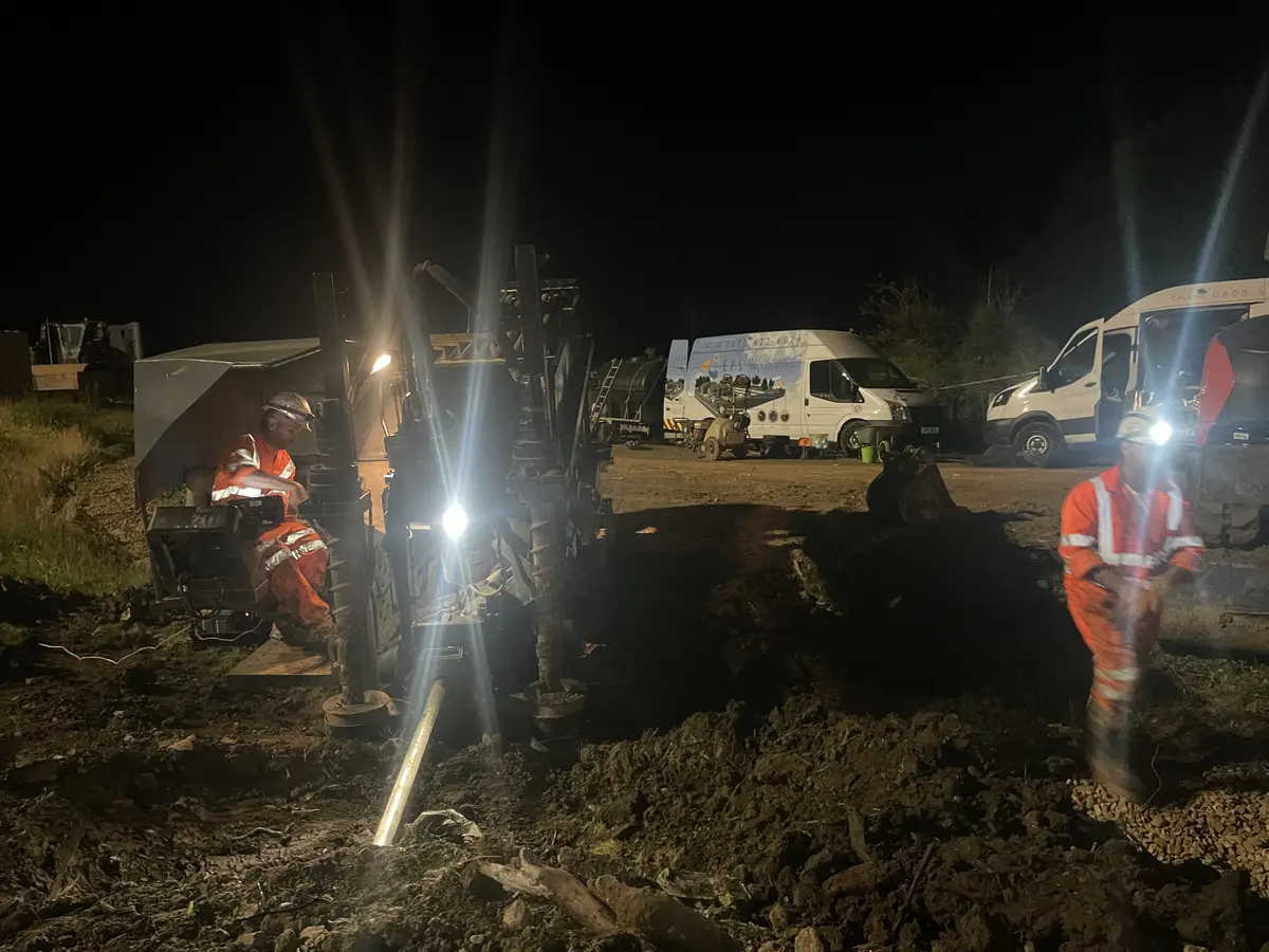 An operator seated at the controls of a directional drilling rig at night under work lights, with the drill string entering the ground and support vans parked behind.