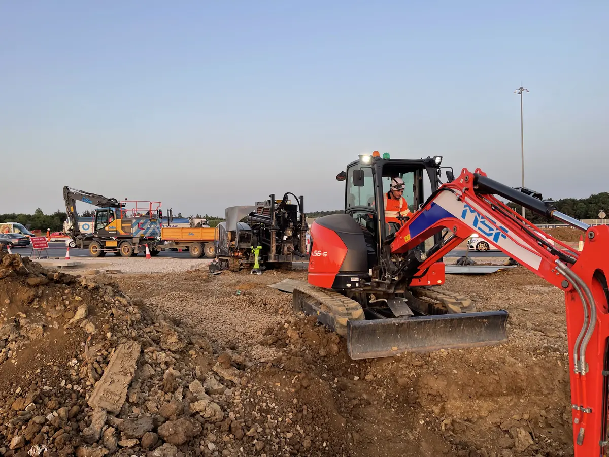 A tracked directional drilling rig and a red MSF mini excavator working alongside each other on made ground, with tipper lorries and plant on the road behind.