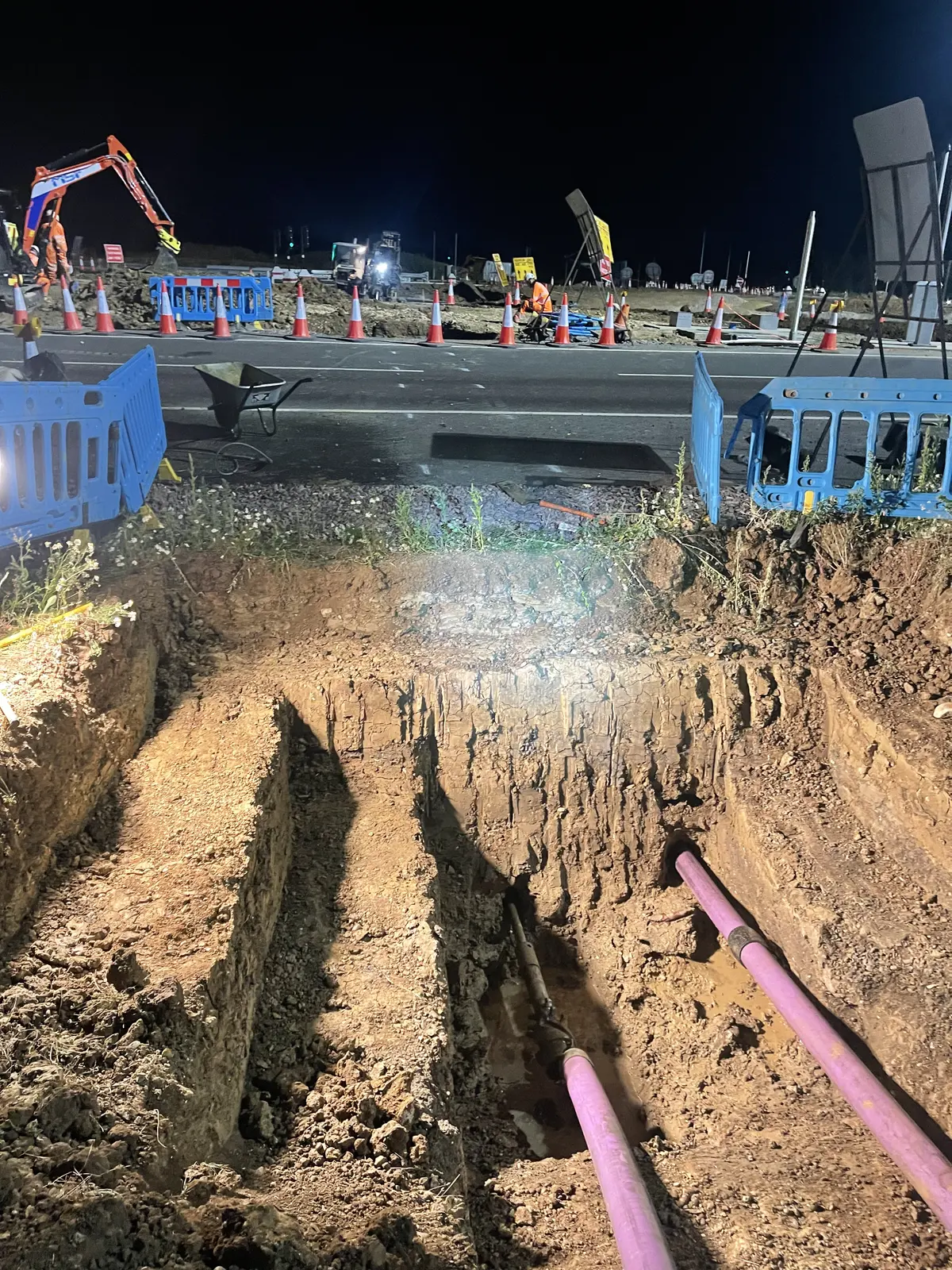 Two purple ducts running through the base of a deep excavation at night, with the carriageway, blue barriers, cones and an excavator visible beyond the verge.