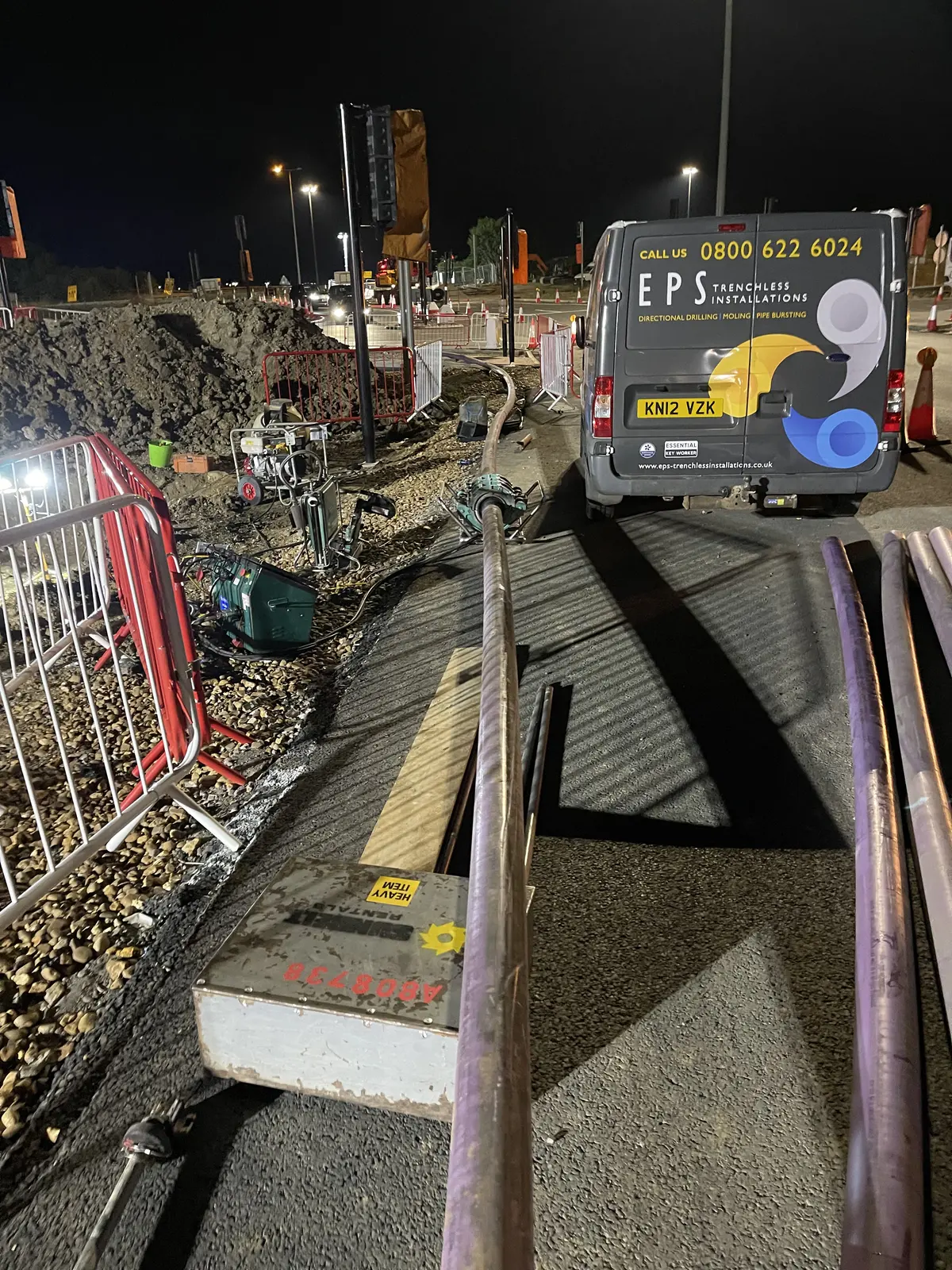 Lengths of purple duct laid out along a road surface at night beside an EPS Trenchless Installations van, with the excavation, barriers and mixing plant behind.