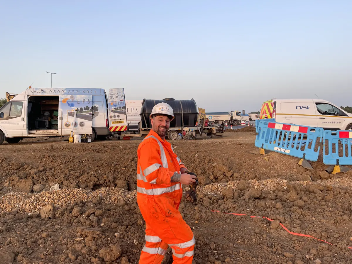 An EPS operative in orange coveralls and a white EPS hard hat, smiling towards the camera on excavated ground, with two EPS vans, a tank trailer and an MSF van behind him in evening light.