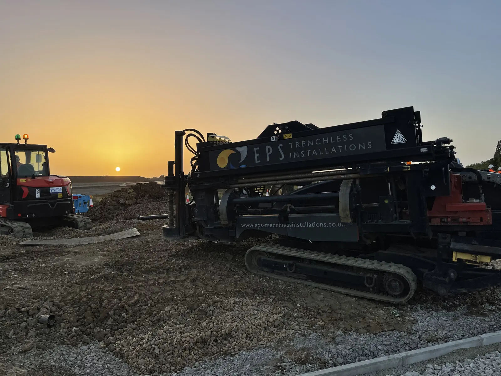 An EPS Trenchless Installations directional drilling rig loaded with drill rods, tracked into position on hardcore beside a red MSF excavator, with the sun low on the horizon behind it.