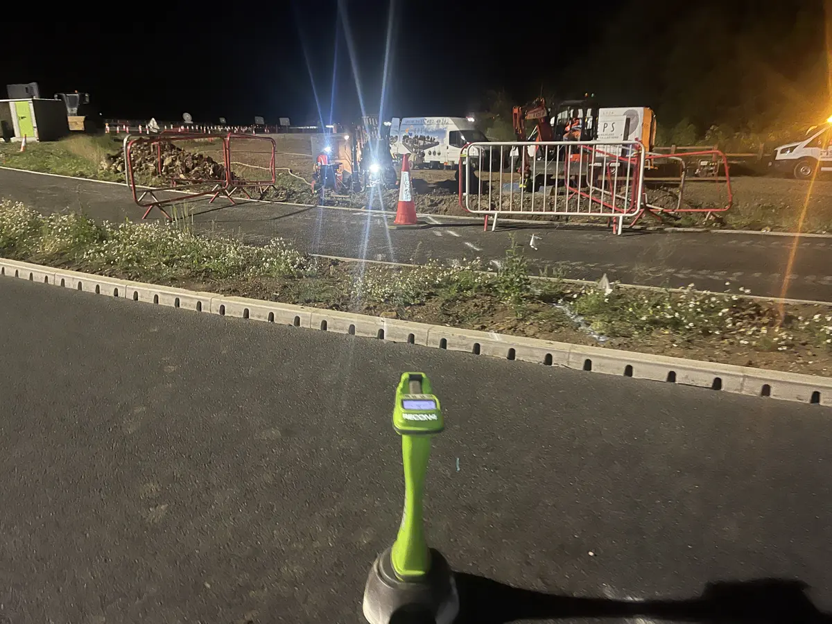 A green Subsite hand-held locator standing upright on the carriageway at night, with the barriered works area, an excavator and site lighting across the kerb line beyond.
