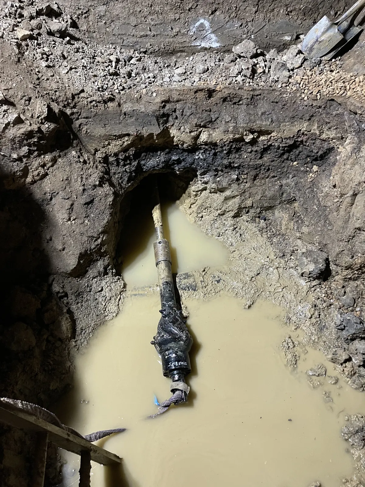 Close view of a reamer assembly attached to the drill string where it enters the bore in an excavation face, above standing muddy water at the base of the pit.