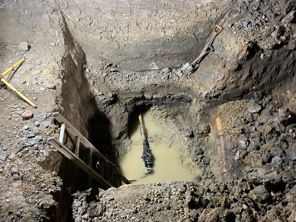 A drill rod and reamer emerging from a bore into a deep excavation part filled with muddy water, with a timber ladder, shovel and survey tripod at the pit edge.