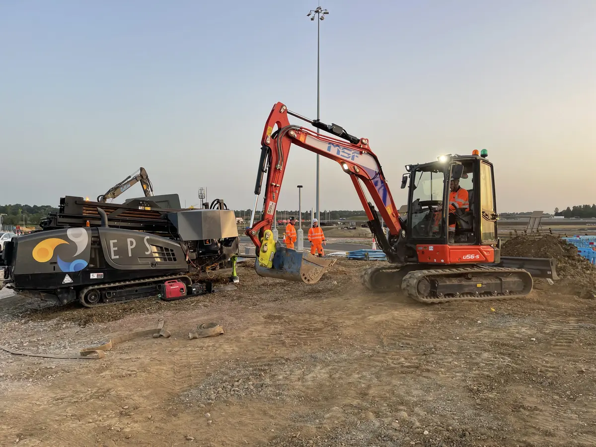A directional drilling rig standing on hardcore beside a newly kerbed carriageway at sunset, with traffic cones and barriers around the works.