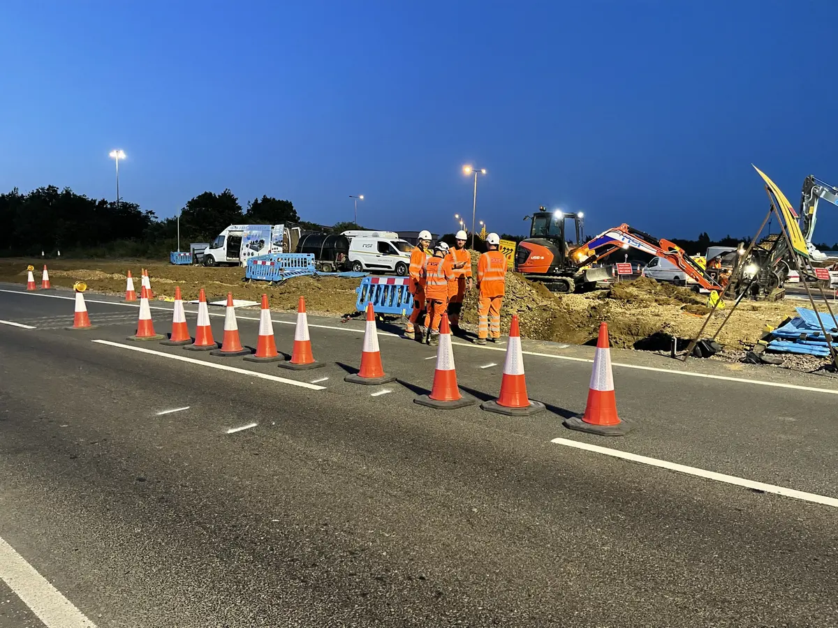 Four operatives in orange hi-vis and white hard hats standing in discussion beside a coned-off works area at dusk, with vans and a tracked excavator behind them.