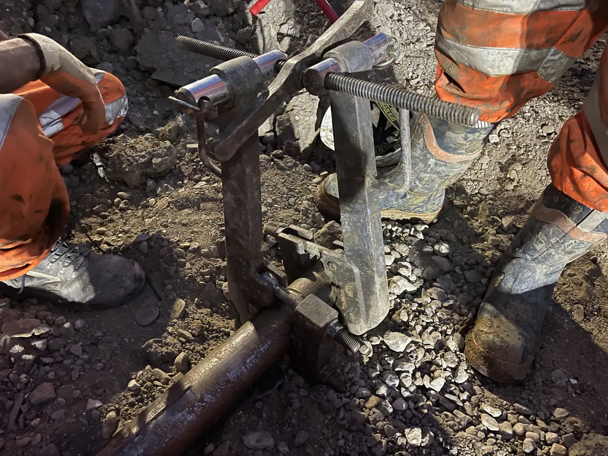 Two operatives in muddy orange hi-vis crouched over a pair of steel breakout wrenches clamped either side of a joint in the drill string, at the stone base of an excavation under work lights at night.