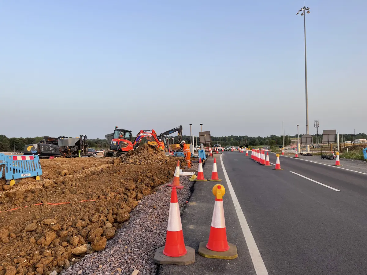 A long line of traffic cones tapering along both edges of a carriageway to close off the working area, with excavated verge, a drilling rig and an excavator beyond.